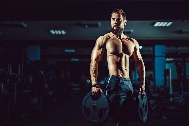 Athlete muscular bodybuilder man posing with dumbbells in gym.