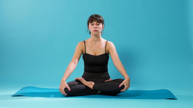 スタジオでのトレーニング活動の後、蓮華座で回復しているアスリート。青い背景の上に体の筋肉を伸ばすためにヨガの練習をしている穏やかな人、フロアマットに座っている瞑想的な女性