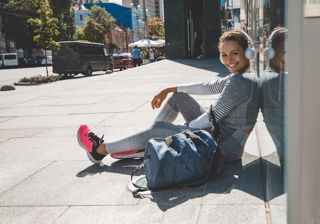 Photo athlete sits with hback against a glass wall, listens to music and smiles