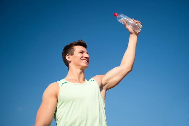 Atleta sorridente in maglietta verde su cielo blu. uomo con bottiglia d'acqua in mani muscolose. stile di vita sano e salute. concetto di sport e fitness. culturista con muscoli bicipiti, tricipiti in una giornata di sole.