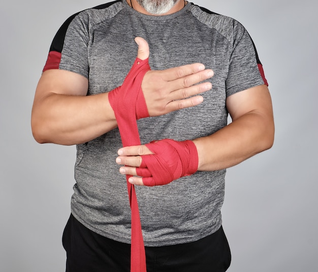 Athlete stand in gray clothes and wrap his hands in red textile elastic bandage 