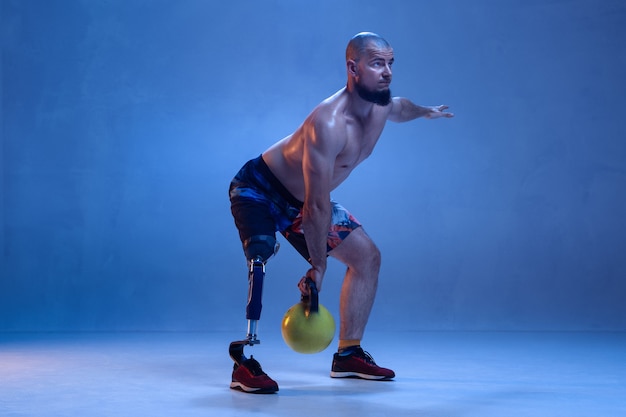 Athlete with disabilities or amputee isolated on blue wall. professional male sportsman with leg prosthesis training with weights in neon. disabled sport and overcoming, wellness concept.