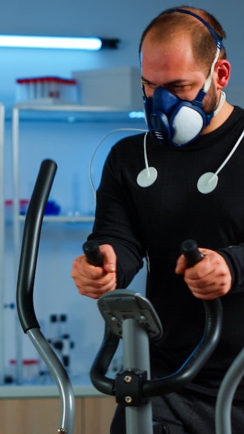 Foto atleta con maschera ed elettrodi attaccati al corpo che corre su cross trainer nel laboratorio sportivo scientifico che monitora lo stato di salute