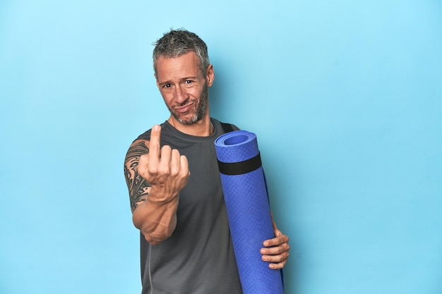 Athlete with a mat on a blue studio backdrop pointing with finger at you as if inviting come closer