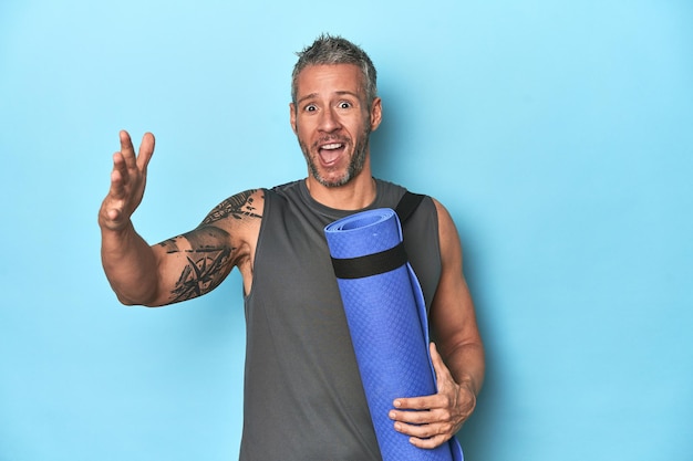 Athlete with a mat on a blue studio backdrop receiving a pleasant surprise excited and raising hands