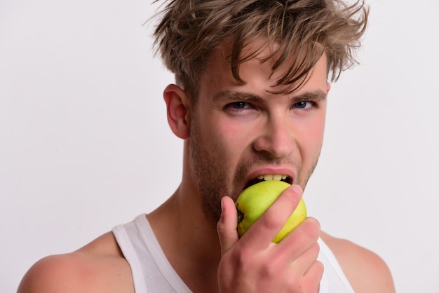 Athlete with messy hair eats fresh fruit Man with green apple in his hand bites it Guy with busy face isolated on light grey background close up Healthy nutrition concept