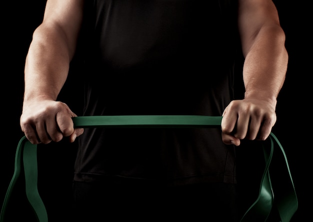 Athlete with a muscular body in black clothes is doing physical exercises with green rubber