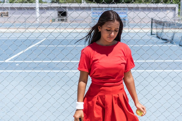 Athlete with racket and ball standing near tennis court fence during break in training