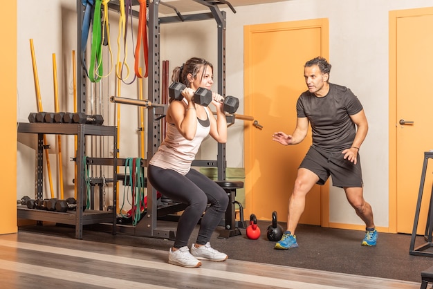 Athlete woman doing squats with dumbbells in gym with help of trainer. Concept of exercise with equipment in gym.