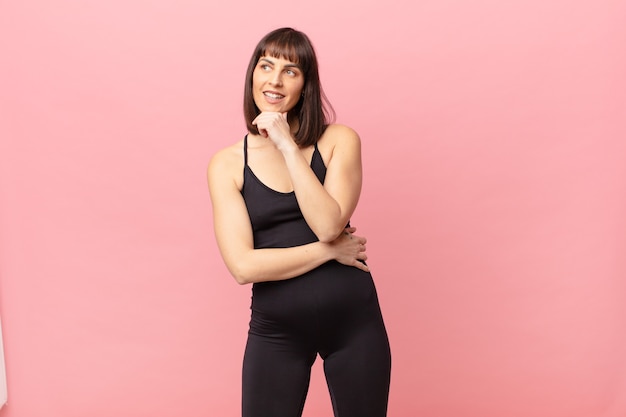 athlete woman smiling with a happy, confident expression with hand on chin, wondering and looking to the side