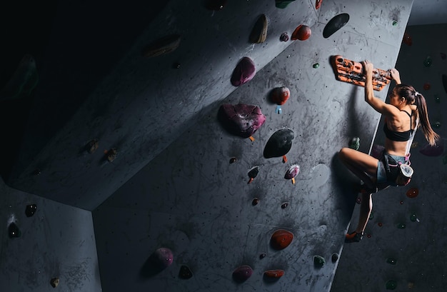 Atleta giovane donna in pantaloncini corti e reggiseno sportivo che si esercita su una parete boulder al chiuso.