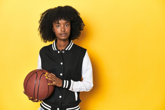 Photo athletic africanamerican teenage girl holding a basketball on a yellow studio backdrop