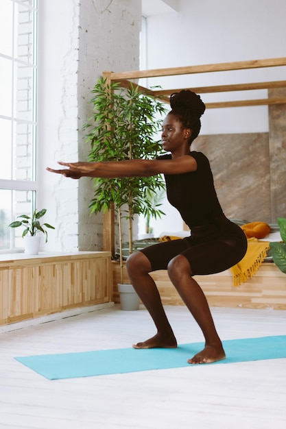 Athletic afro american woman squatting with arms extended forward side view training on gymnastic mat in decorated studio Sports course exercise technique fitness keeping body fit