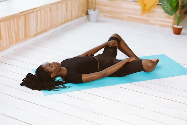 Athletic afro american woman with dreadlocks lying on back and stretching leg muscles training on gym mat in light studio sports course exercise technique body stretching gymnastic exercise