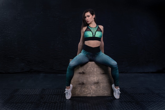 Photo athletic athlete dressed in fitness clothing poses on a dark background squatting on a wooden box