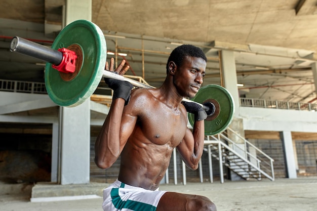 Athletic black young man lifting a heavyweight barbell in outdoor gym under the bridge Healthy lifestyle concept
