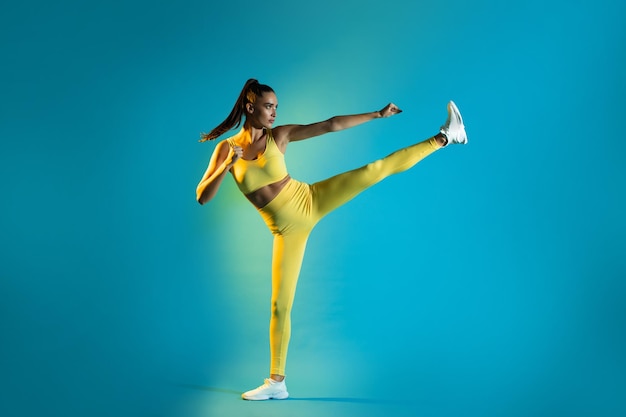 Athletic female doing karate side kick clenching fists blue background