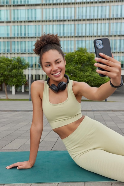 Athletic female model dressed in sportswear sits on fitness mat winks eyes takes selfie rests after doing physical exercises wears headphones around neck poses against urban building outdoors