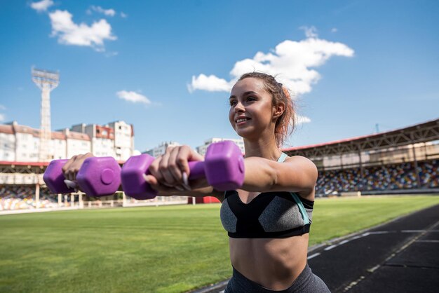 シティスタジアムでダンベルを使ってトレーニングをしているスポーツウェアのアスレチック女性トレーナー