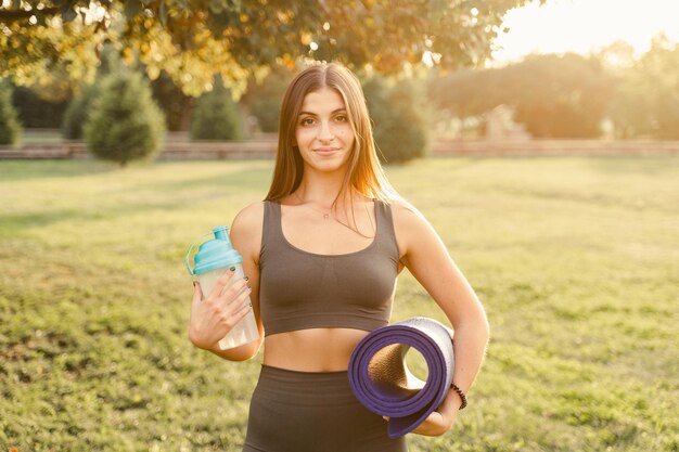 Спортивная девушка с тренировочным ковриком и бутылкой воды на открытом воздухе Реклама магазина спортивного питания и снаряжения