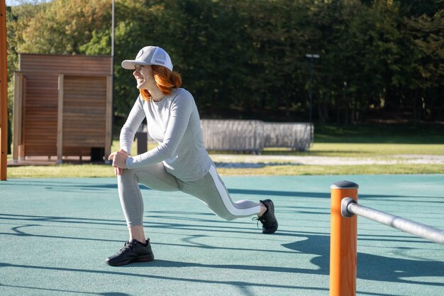 Athletic ginger woman warming up for morning training outdoor on urban sports ground in autumn sunny day.