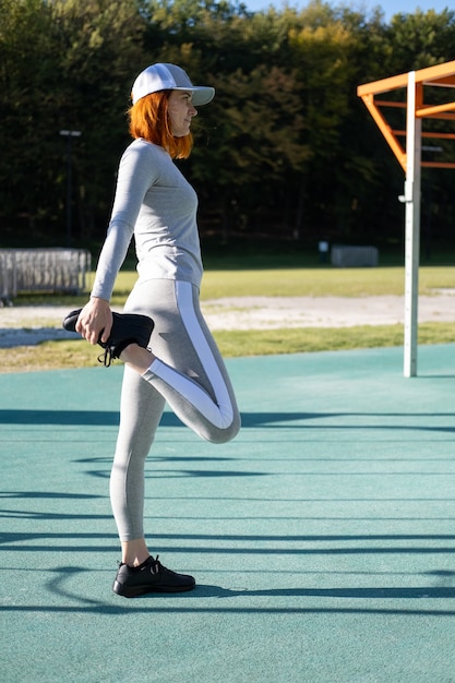 Athletic ginger woman warming up for morning training outdoor on urban sports ground in autumn sunny day.