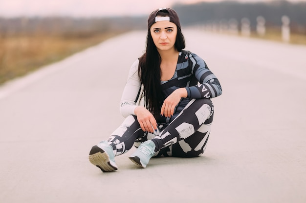 Athletic girl in sportswear with brutal look sitting on the asphalt