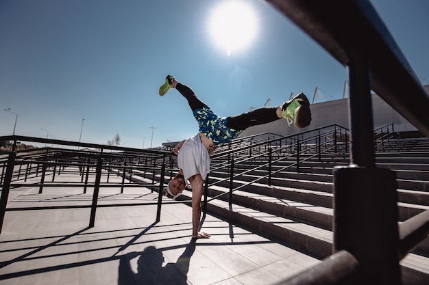 Athletic guy with headband on his head dressed in the white t-shirt, black leggings and blue shorts is doing the trick standing on one hand on the stairs next to railing outside on a sunny day .