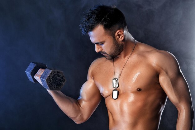 Спортивный (ый) красивый мускулистый мужчина делает упражнения с гантелями. Сильный культурист с голым торсом на темном фоне