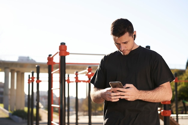 Athletic man in a bar park checking his phone. He is wearing a wireless headset.