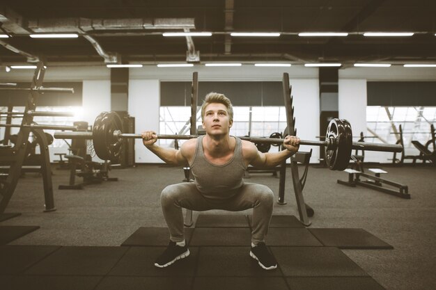 Athletic man exercising with barbell at gym. Athletic man in sportswear doing squats with barbell on his shoulders in gym.