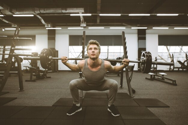 Uomo atletico che si esercita con il bilanciere alla palestra. uomo atletico in abiti sportivi facendo squat con bilanciere sulle spalle in palestra.