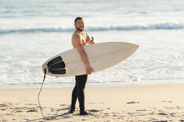 An athletic man surfer with naked torso standing by the sea and showing a thumb up looking in the ca