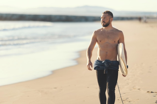 Foto un surfista uomo atletico con il torso nudo che cammina sulla riva del mare