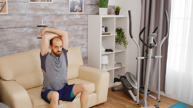 Athletic man training triceps muscle with dumbbell sitting on sofa in living room.