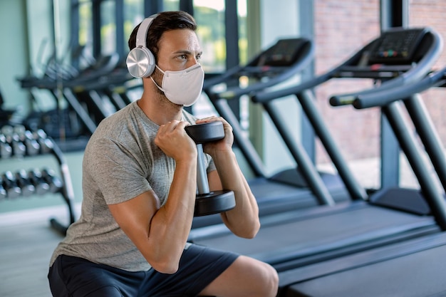 Athletic man wearing face mask while using dumbbell and exercising squats in a gym