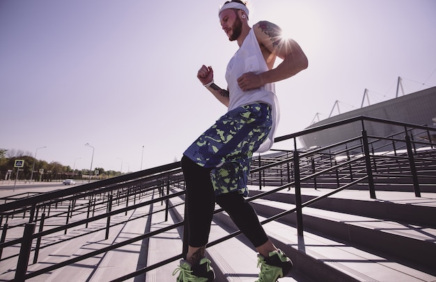 Uomo atletico con fascia in testa vestito con maglietta bianca, leggings neri e pantaloncini blu sta correndo giù per le scale fuori in una giornata di sole.