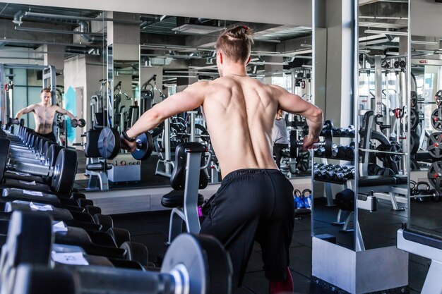 Athletic man with a naked torso in the gym lifts dumbbells. Photo from the back