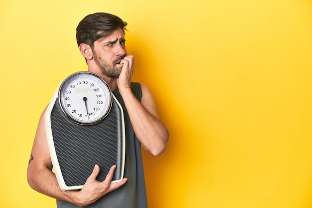 Athletic man with scale on a yellow studio backdrop biting fingernails nervous and very anxious