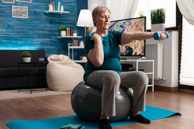 Athletic pensioner in sportswear watching online aerobic workout using tablet sitting on swiss ball ...