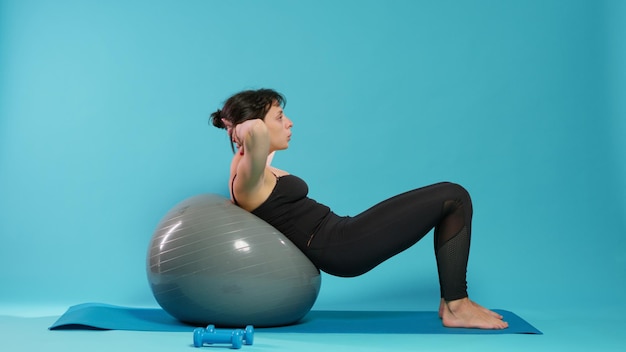 Athletic person doing physical effort on fitness toning ball, training body muscles. Fit woman using sport equipment to do gymnastics exercise and endurance activity over background.