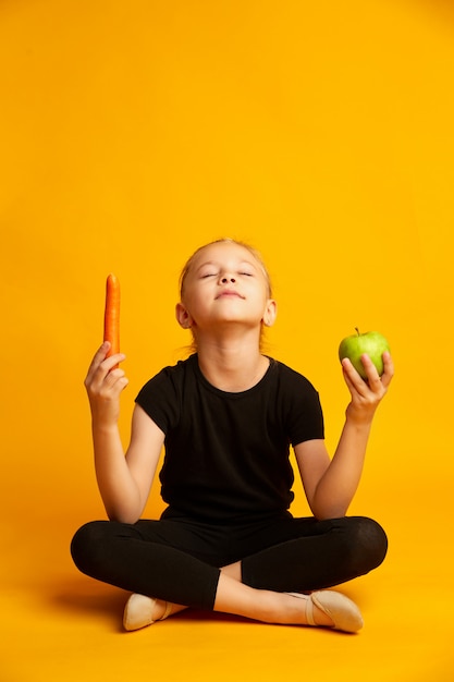 Спортивная семилетняя девочка держит зеленое яблоко и морковь в руках, изолированных на желтом
