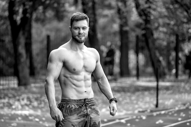 スポーツヤードで屋外でポーズをとるアスレチック上半身裸の男性フィットネスモデル素敵な筋肉の体ハーフワイスト写真カメラを見てクローズアップ