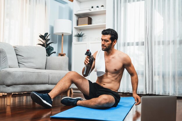 Athletic and sporty man drinking water on fitness mat at gaiety home