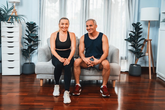 Athletic and sporty senior couple portrait in sportswear sitting on sofa as home exercise concept Healthy fit body lifestyle after retirement Clout