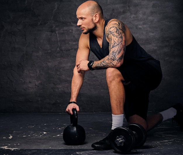 Athletic tattooed, shaved head sporty male standing in knee and holds Kettlebell.