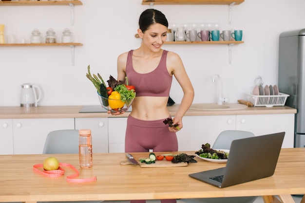 Athletic woman blogger nutritionist prepare a salad with fresh vegetables and conducts a video conference on healthy eating on laptop in the kitchen