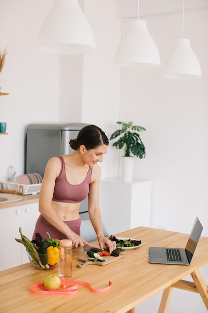 Athletic woman blogger nutritionist prepare a salad with fresh vegetables and conducts a video conference on healthy eating on laptop in the kitchen