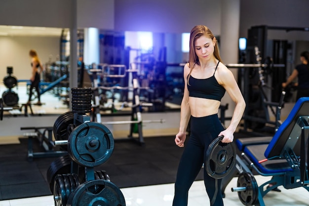 Athletic woman holds a black pancake rod and looks at camera in\
the gym in the sport equipment background health life concept