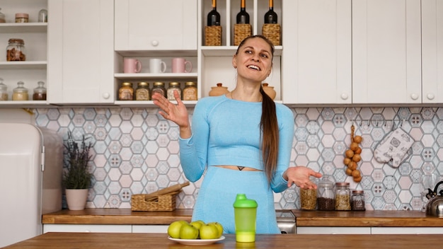 La donna atletica parla di mangiare sano in cucina e ride. sul tavolo ci sono mele verdi e una bottiglia verde per una bevanda sportiva e acqua